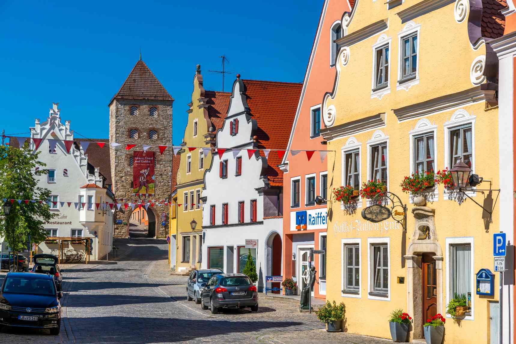 Historic street in Rothenburg ob der Tauber, Germany, featuring colorful medieval buildings, flower-decorated windows, cobblestone streets, and a historic stone gate tower along Germany’s Romantic Road