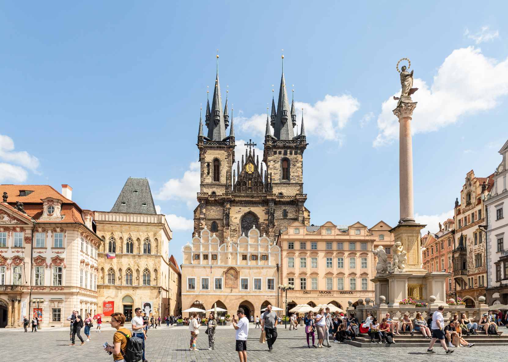 Prague Old Town Square in daylight, featuring the Gothic twin towers of the Church of Our Lady before Týn, colorful historic buildings, and the Marian Column