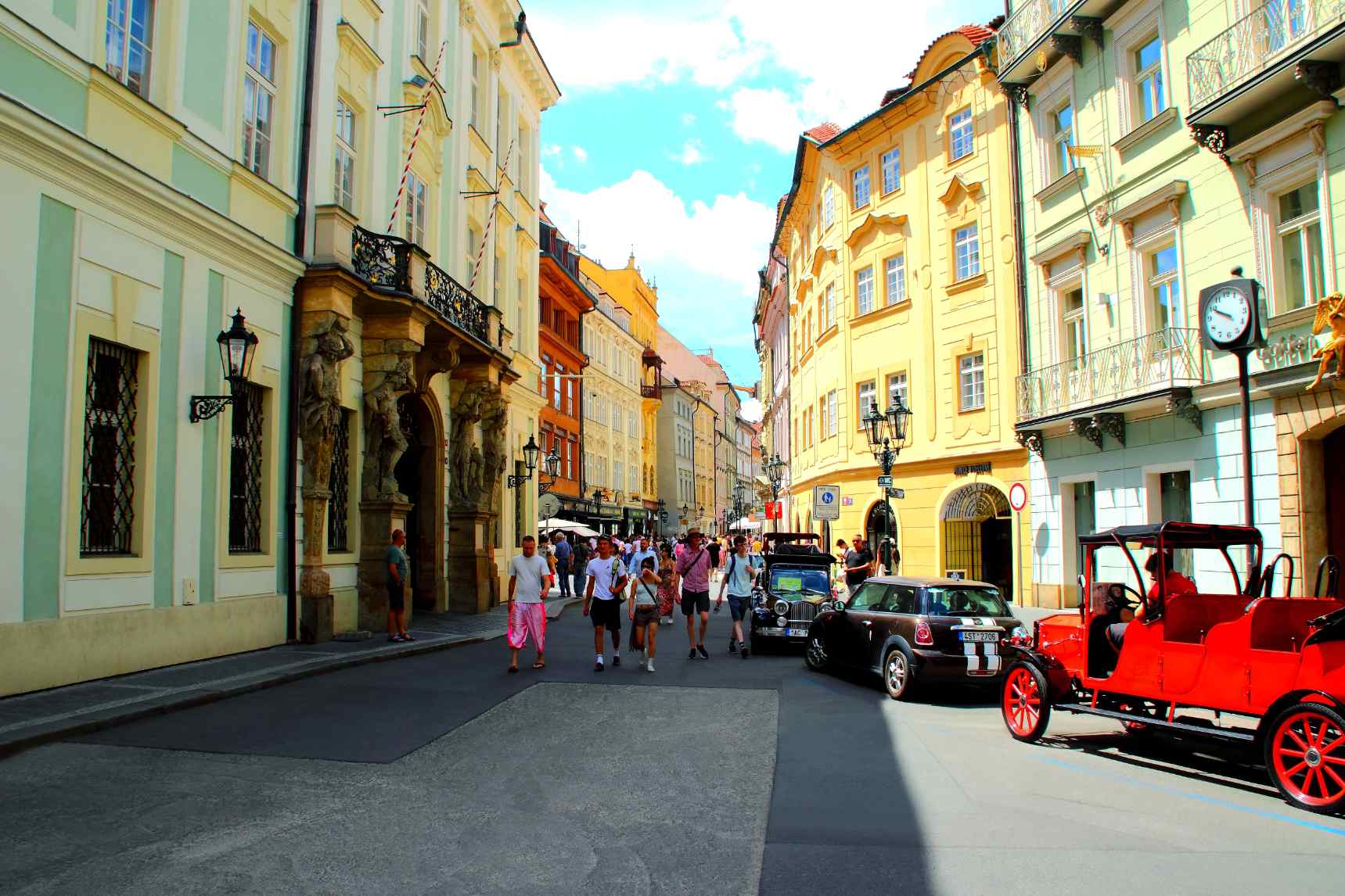 Historic street in Prague, Czech Republic, featuring colorful European architecture, ornate building facades, sightseeing visitors, and a red vintage tourist vehicle