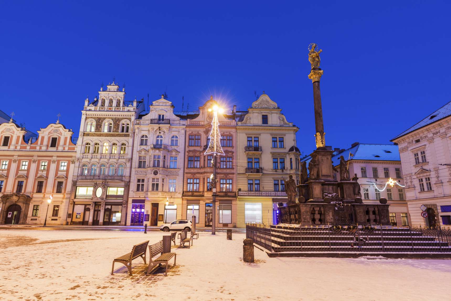 Snow-covered Republic Square in Pilsen, Czech Republic, illuminated historic buildings and the Marian Plague Column with a golden statue on a winter evening