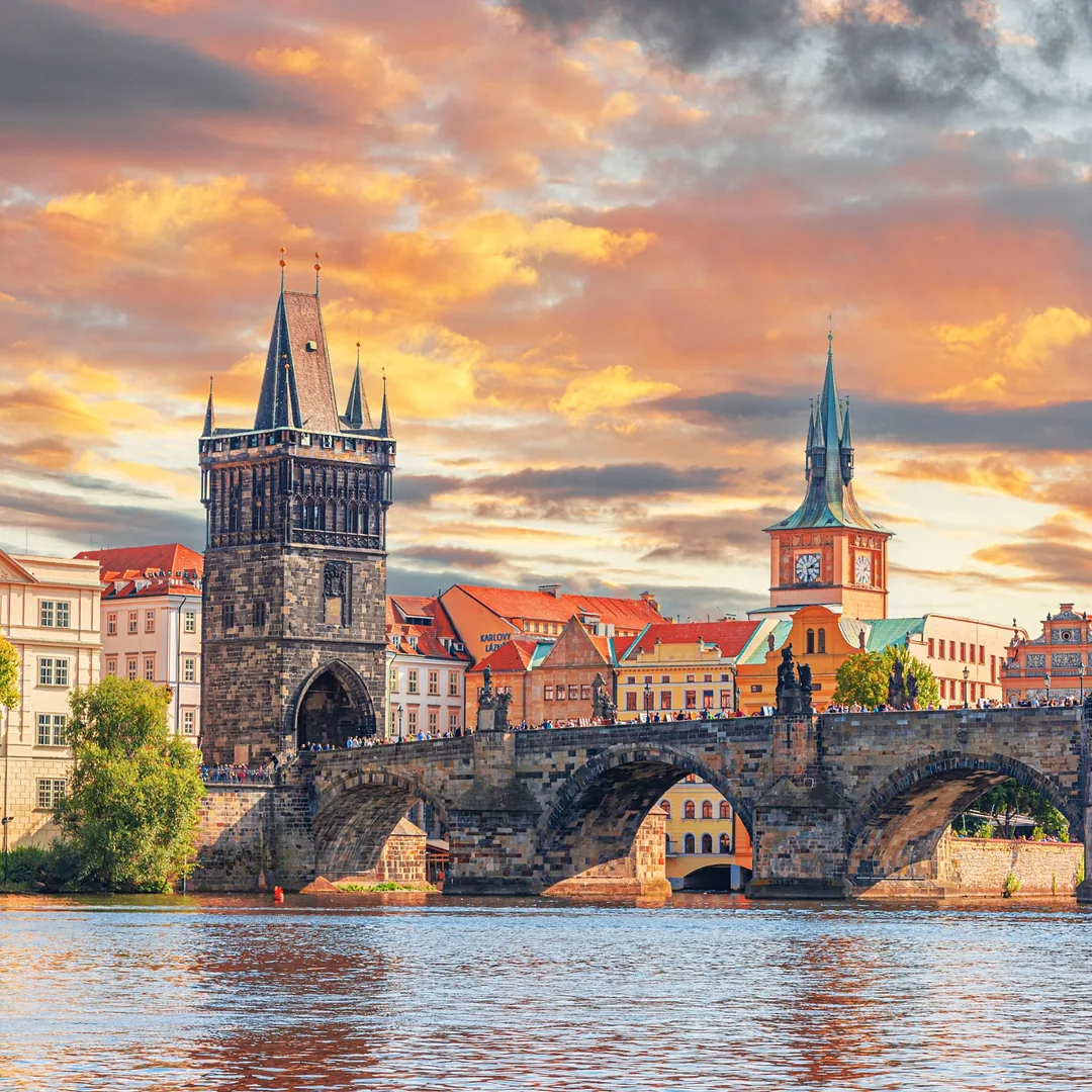 Charles Bridge in Prague at sunset, spanning the Vltava River with its historic Gothic bridge tower, red-roofed Old Town buildings, and colorful evening sky
